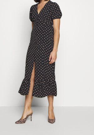 Femme portant une robe midi noire à pois roses, manches courtes bouffantes, fente à l'avant, et des chaussures à talons hauts imprimé léopard.