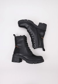Botas de cuero negro hasta el tobillo con un tacón grueso, que presentan cordones en la parte frontal, un zipper lateral y un detalle de costura naranja.