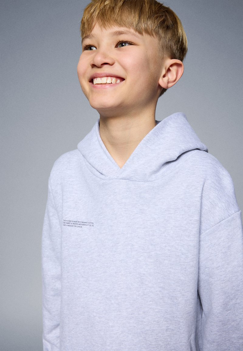 Light gray hoodie made of organic cotton, featuring a large hood, dropped shoulders, and small printed text on the chest. Soft texture.