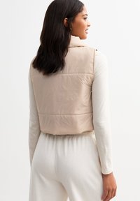 Beige sleeveless puffer vest with a high collar, featuring a shiny texture and vertical quilting, paired with a light cream long-sleeve top.