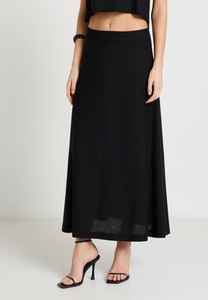 Frau trägt einen langen schwarzen Rock, ein kurzes schwarzes Oberteil, schwarze Sandalen mit hohen Absätzen und ein schwarzes Armband am linken Handgelenk.