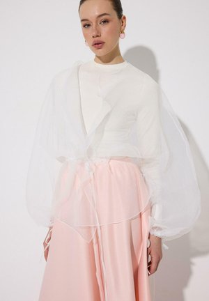 Naine, kes kannab valget läbipaistvat sädeliste varrukatega pluusit valge pika varrukaga särk ja voolavat heledat roosat seelikut, roosade kõrvarõngastega valgel taustal.