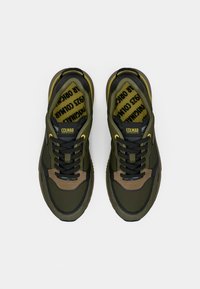 Adidași verzi închiși cu o suprafață texturată, șireturi negre și accente maro. Au o talpă interioară cu un model și cu branding.