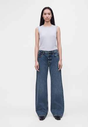 Femme aux longs cheveux foncés portant un haut blanc sans manches et un jean bleu à jambes larges, debout devant un fond clair uni.
