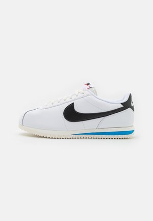 Valkoinen Nike Cortez -tennari mustalla swoosh-logolla, sinisellä raidoituksella pohjassa, valkoisilla nauhoilla ja valkoisella välipohjalla yksinkertaisella taustalla.
