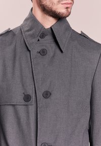Szary płaszcz trencz z usztywnionym kołnierzem, podwójnym zapięciem na guziki oraz pagonami na ramionach. Wykonany z teksturowanej tkaniny o gładkim wykończeniu.