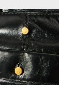 Fekete bőr kézitáska közeli kép, jellegzetes arany hardver gombokkal és részletes varrással, sima tapintással és letisztult dizájnnal.