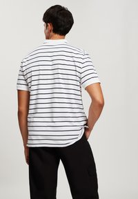 Biała bawełniana koszulka z krótkim rękawem, ozdobiona granatowymi poziomymi paskami. Posiada klasyczny kołnierzyk oraz luźny krój, z nieco dłuższym tylnym zakończeniem.