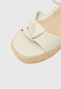Kreminės spalvos espadrilės sandalai su minkštu audinio mazgu, lygiu viršumi ir audiniu pintu jūrinės šiaudų platforminiu padu su tekstūruota detalizacija.
