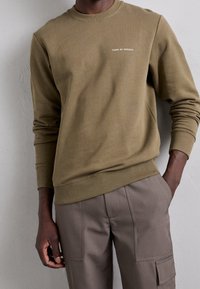 Man i olivgrön sweatshirt med "Tiger of Sweden"-logotyp och taupe cargobyxor, hand i fickan, stående mot en grå vägg.