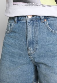 Primer plano de la parte media usando jeans de mezclilla azul claro y una camisa blanca con texto negro parcialmente visible sobre la cinturilla.