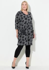 Schwarz-weiße Tunika mit Muster und Drei-Viertel-Ärmeln, mit zwei Taschen, kombiniert mit schwarzen Leggings und spitzen Absätzen.