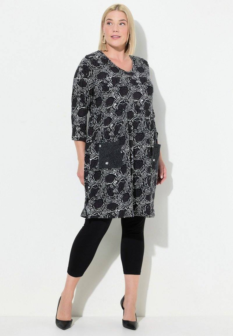 Schwarz-weiße Tunika mit Muster und Drei-Viertel-Ärmeln, mit zwei Taschen, kombiniert mit schwarzen Leggings und spitzen Absätzen.