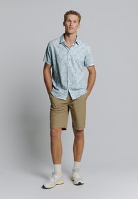 Korte mouwen lichtblauw shirt met bloemenpatroon, gecombineerd met beige shorts. Witte sneakers met grijze accenten en lichte sokken.