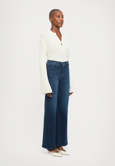 Donna con capelli corti, che indossa un maglione bianco a maniche lunghe a coste, jeans scuri a vita alta con gamba svasata e décolleté bianchi a punta.