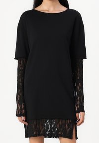 Zwarte top met korte mouwen en lange kantmouwen; de kanten zoom onthult een patroon. Zacht materiaal, relaxte pasvorm met zijsplitten.