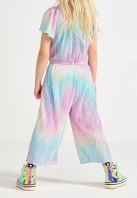 Bambino con lunghi capelli biondi che indossa una tuta plissettata arcobaleno pastello e sneaker alte iridescenti lucide, in piedi su uno sfondo bianco.