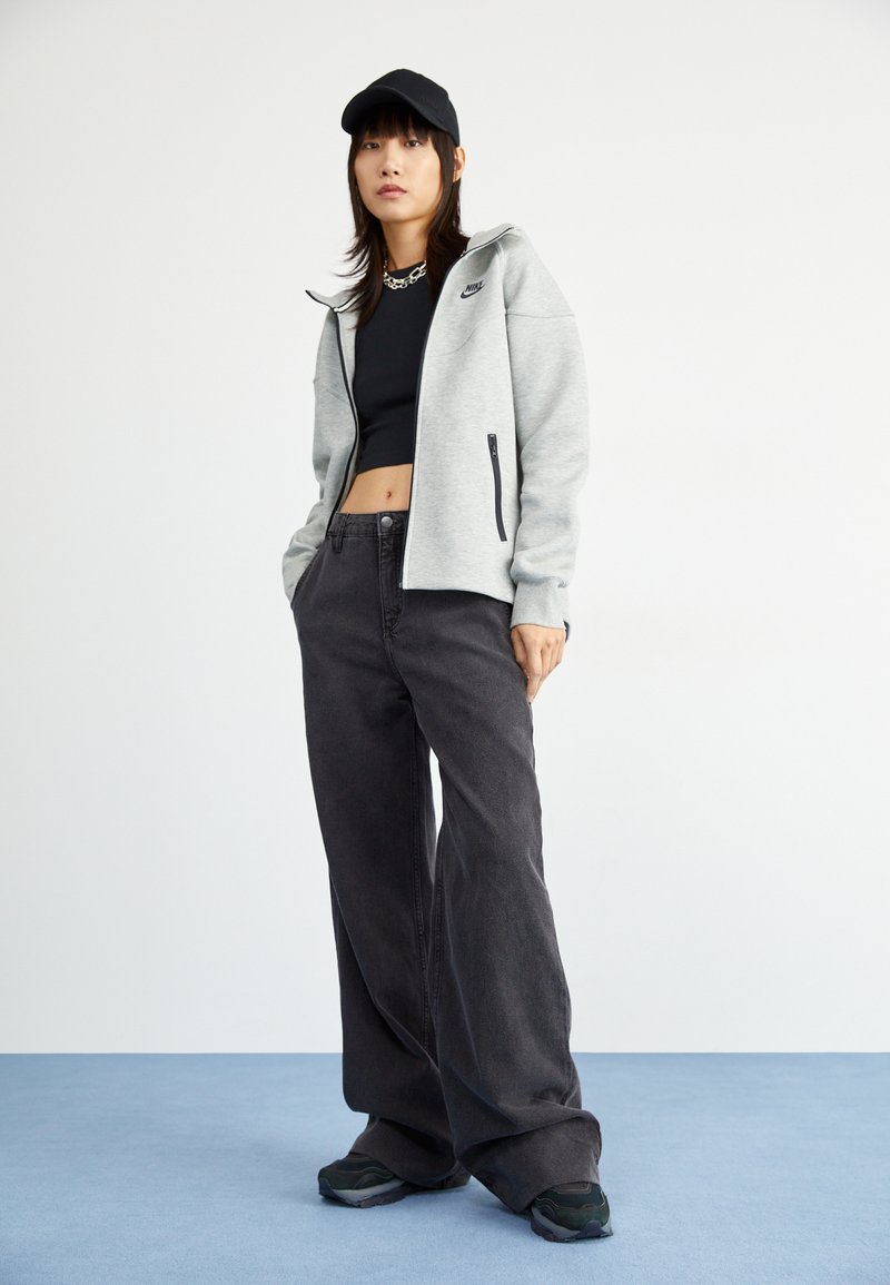 Grauer Reißverschluss-Hoodie, schwarzes Crop-Top, schwarze weit geschnittene Hose, schwarze Kappe und grüne Sneakers, stehen vor einem hellen Hintergrund.