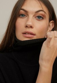 Svart ribbad polotröja med hög krage. Mjuk textur, kroppsnära design. Modellen håller kragen nära ansiktet.