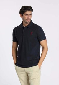 Polo shirt en coton noir avec un col à boutons et des manches courtes, arborant un logo rouge brodé sur la poitrine gauche.
