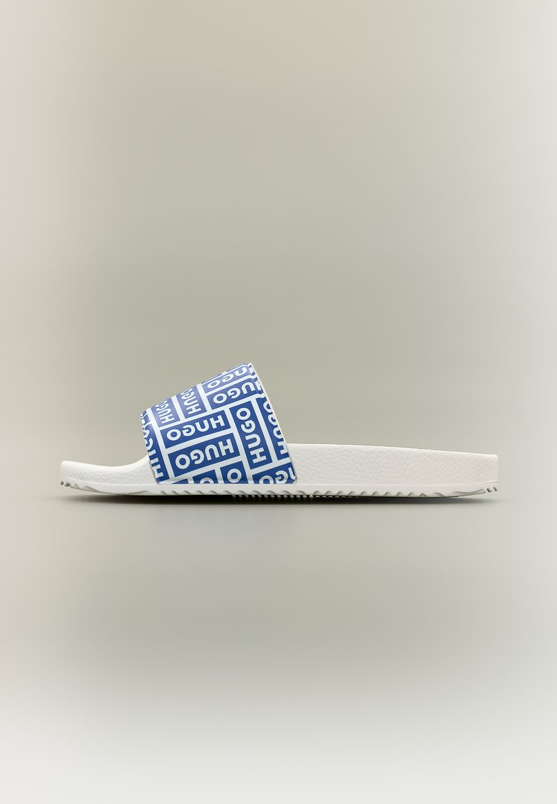 Sandale blanche à semelle plate avec une bride bleue siglée, affichant le texte répété "HUGO" en lettres blanches majuscules, vue de profil sur un fond uni.