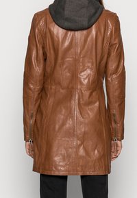 Bruine leren jas met een getailleerd design, capuchon van donkere stof, zichtbare stiksels en ritszakken aan de zijkant. Gladde textuur, knielang.