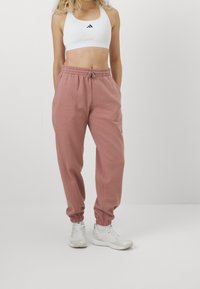 Pantaloni della tuta rosa in cotone con polsini elasticizzati e cintura regolabile, abbinati a un reggiseno sportivo bianco con logo nero. Scarpe da ginnastica bianche.