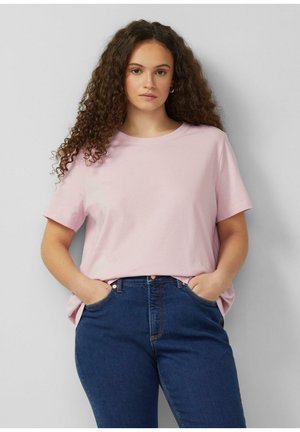 Młoda kobieta o długich kręconych włosach, ubrana w jasnoróżowy t-shirt z krótkim rękawem i ciemnoniebieskie dżinsy, stojąca z rękami w kieszeniach.