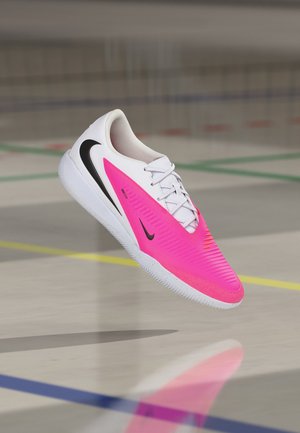 Hvid og pink Nike indendørs fodboldstøvle med sort swoosh-logo svævende over et gulv i et motionscenter med banemarkeringer.