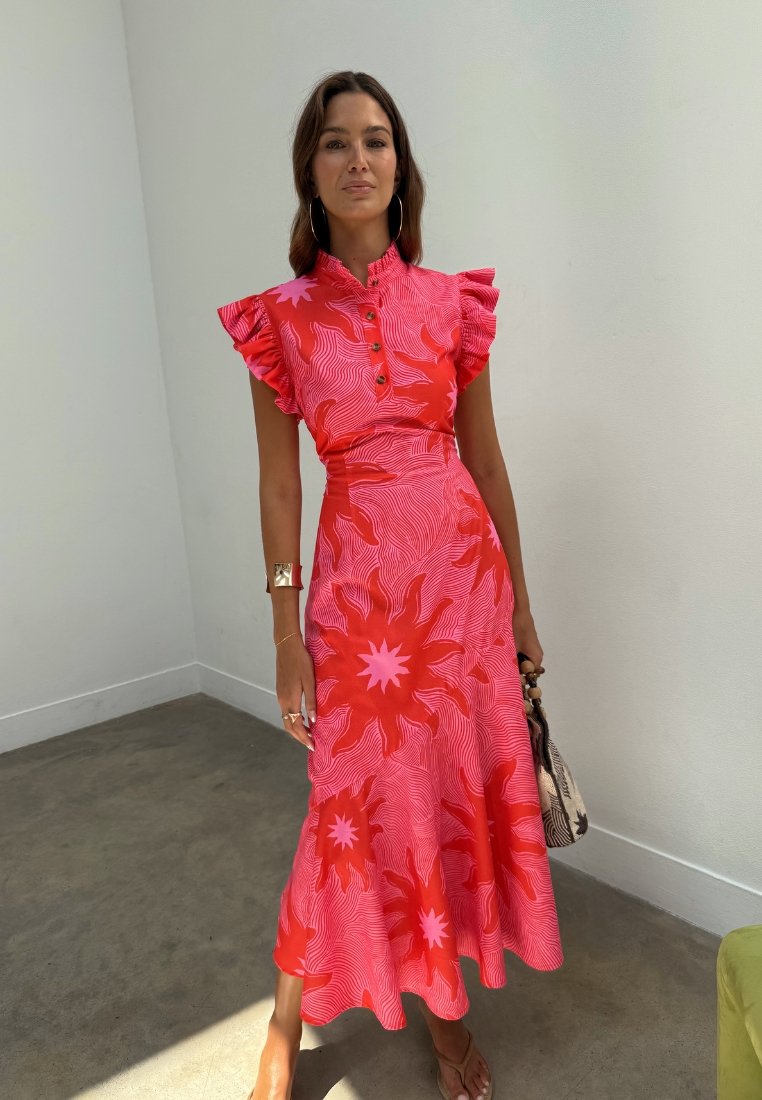 Femme portant une robe midi rouge et rose à motif de soleil avec des manches à volants, tenant un petit sac à main, debout dans une pièce minimaliste.