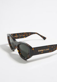 Gafas de sol en carey negro y marrón con lentes grises, que presentan un diseño audaz y angular, y la marca "BIMBA Y LOLA" en la patilla.