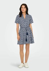 Kratka haljina s kratkim rukavima u mornarsko plavom i bijelom cvjetnom uzorku, sa ovratnikom, pojasom u struku i lepršavom suknjom, uparena s bijelim tenisicama.