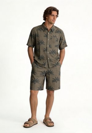 Mand stående med hænderne i lommerne iført matchende olivengrøn kortærmet skjorte og shorts med sorte palme træer og beige sandaler.