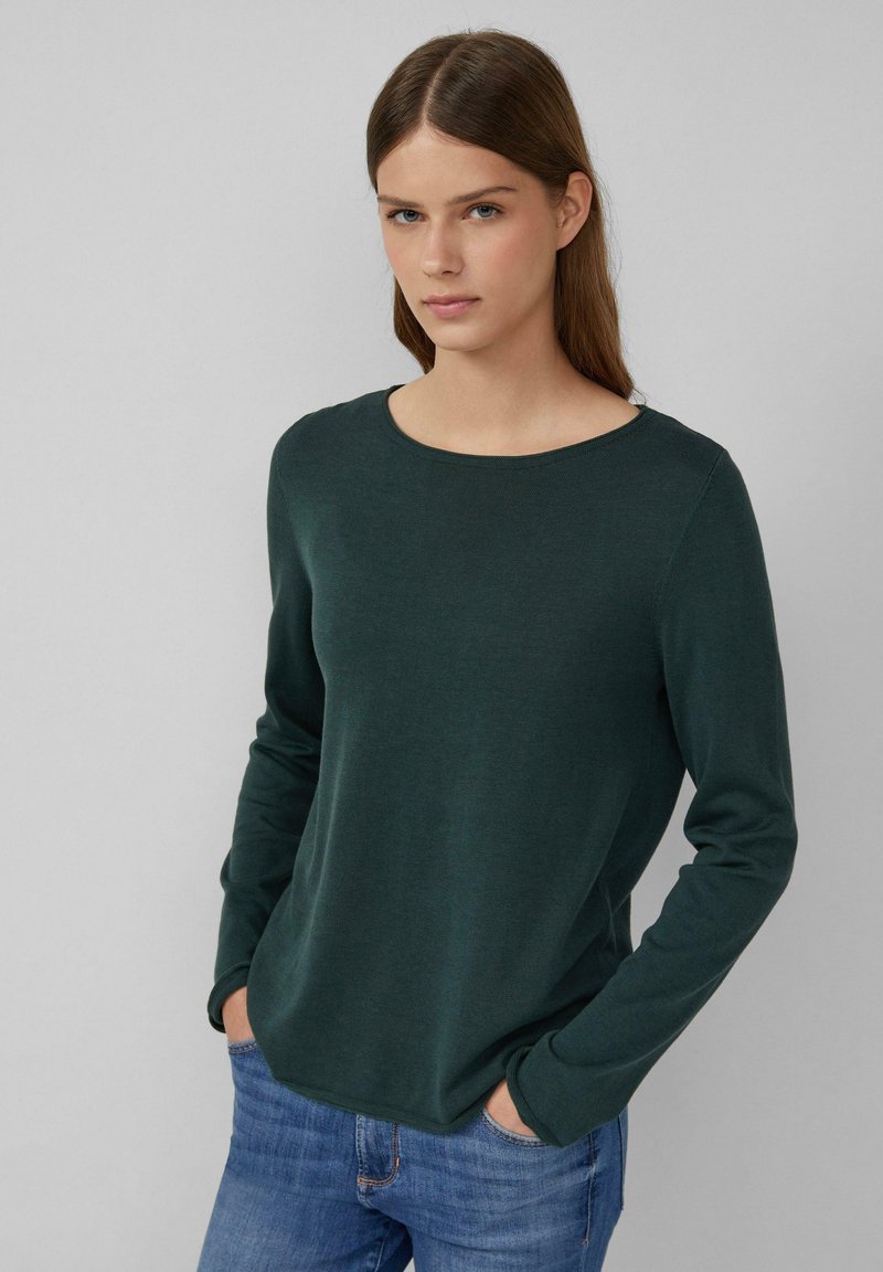 Dunkelgrüner Langarmpullover mit rundem Halsausschnitt und glatter Textur, kombiniert mit hellblauen Jeans. Einfaches, lässiges Design.