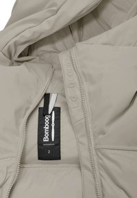 Beige gewatteerde jas met een gladde binnenkant, zichtbare naden en een zwart label met het merk "Bombog" en verzorgingsinstructies.