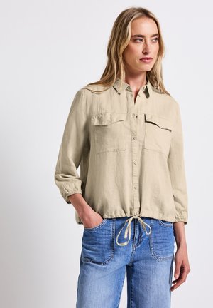 Beige, langwerpige button-up shirt met twee borstzakken, geplooide manchetten en een trekkoordonderrand, gecombineerd met lichtblauwe denim jeans.