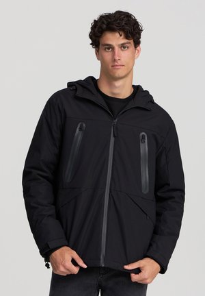 Giacca impermeabile nera con cappuccio, dotata di due tasche sul petto con zip, una texture liscia, polsini elasticizzati e un design dalla vestibilità rilassata.