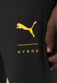 Crne atletske tajice s žutim Puma logotipom i žutim "HYROX" natpisom, izrađene od rastezljivog materijala s glatkom teksturom.