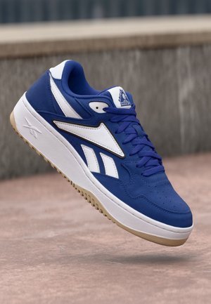 Blue athletic shoe with white accents, textured upper, perforated toe, and a beige rubber sole. Features a branded tongue and lace closures.