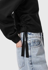 Sudadera negra con lados fruncidos, lazos de ajuste y mangas largas, combinada con pantalones cortos de mezclilla descoloridos en azul claro, con un bolsillo delantero.