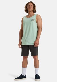 Hellgrünes Tanktop mit quadratischem Taschen-design, kombiniert mit dunkelgrauen Shorts und navy-farbenen Sneakers. Weiße Crew-Socken runden das Outfit ab.