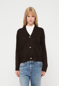 Cardigã tricotado castanho com decote em V, cinco bot�ões grandes e dois bolsos frontais. Bainha e punhos canelados, usado sobre uma blusa branca e jeans azuis.