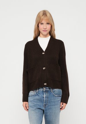 Barna kötött kardigán V-nyakkal, öt nagy gombbal és két elülső zsebbels. Ribbelt szegő és mandzsetta, fehér toppal és kék farmernadrággal viselve.