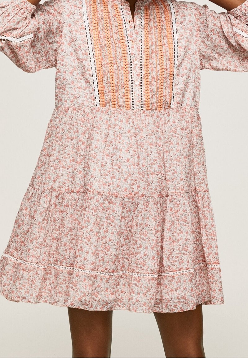 Robe florale rose et blanche avec manches longues, broderie verticale orange sur le corsage et jupe à étages portée par une personne debout.