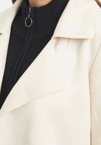 Light beige suede jacket with a wide collar, layered over a ribbed black high-neck top featuring a silver zipper detail.
