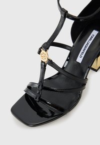 Sandália de salto alto em verniz preto com bico quadrado, apresentando várias tiras estreitas e um detalhe do logótipo dourado na lateral.