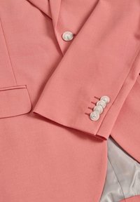 Blazer rosa chiaro con una texture liscia, una tasca e quattro bottoni bianchi sul polsino con dettagli di cucitura sottili. Interno foderato.