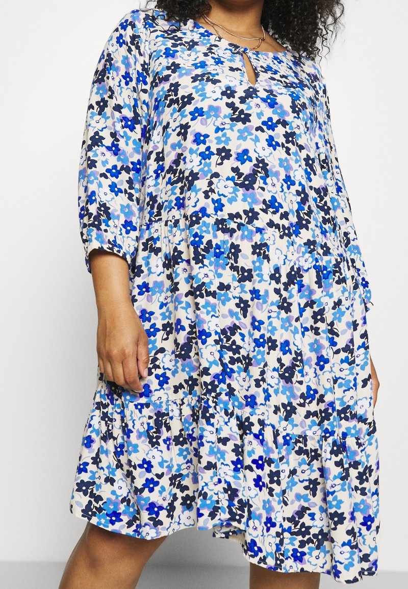 Femme portant une robe ample mi-longue avec un imprimé floral bleu, noir et blanc et des manches trois-quarts.