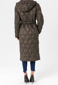 Kviltad lång kappa i mörk olivgrön med huva, insvängd midja och diamantmönster. Matchar med slitna blå jeans och svarta klackar.