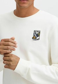 White cotton sweatshirt featuring ribbed neckline and cuffs, with a colorful embroidered patch depicting a foot on a banana and "BORN LUCKY."